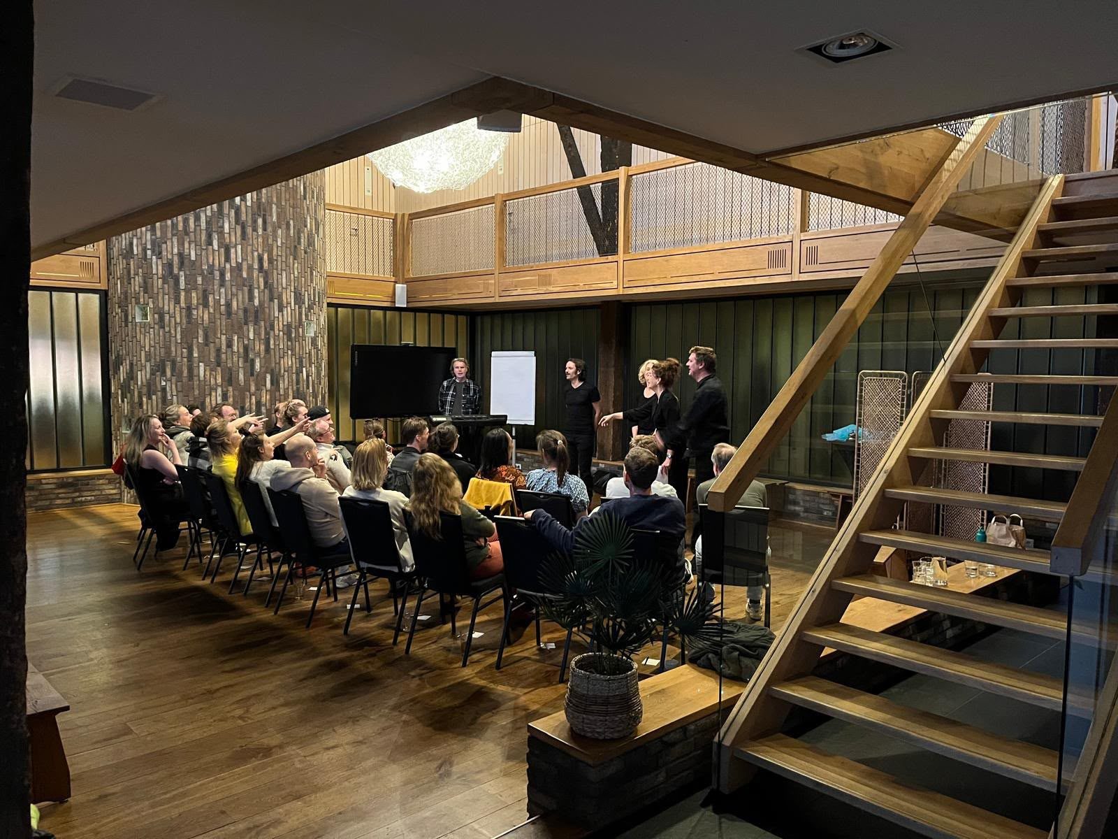 Zaal met publiek op stoelen en spelers voor een groene wand.
