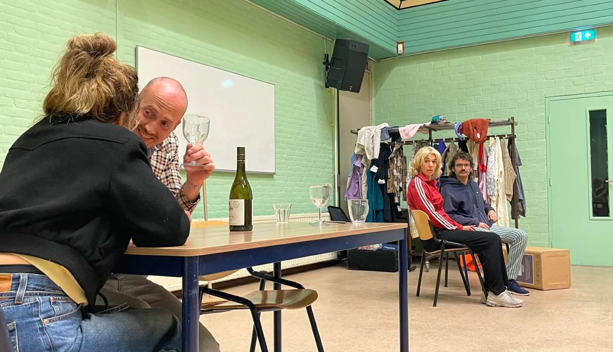 Twee spelers van Theatergroep PIT aan een tafel met een wijnfles tijdens een training in Villa Nova, op de achtergrond kijken medespelers toe bij een rek met kostuums